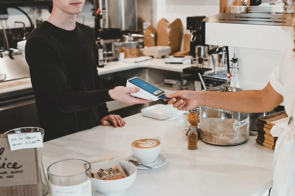 A café employee holds a payment terminal while a customer inserts a card, a latte on the counter.